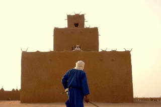 Person in desert looks up at structure. Another person perched on the structure looks down.