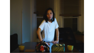 The artist is central in the frame, standing behind a table. The room she is in is dark, though she is lit by some of the TV. On the table is a green ceramic planter pot, a brown envelope, two warpped parcels, and another green ceramic planter pot.