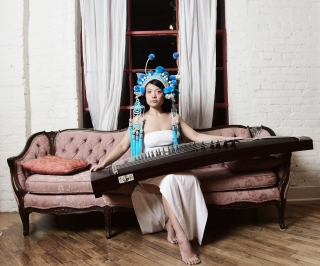 Wu Fei on a Victorian couch, holding an instrument wearing a turquoise headdress..