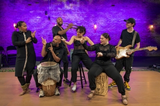 Full band on stage with dancers. Drums, purple background, fun poses.